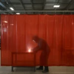 Semitransparent red industrial welding curtain protecting a factory worker from arc radiation, highlighting CSA W117.2 compliance.